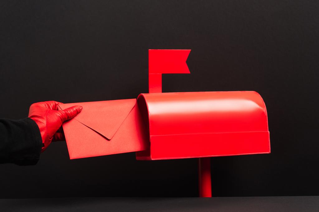 bijgesneden weergave van persoon in rode handschoen zetten envelop in brievenbus op zwart  - Foto, afbeelding