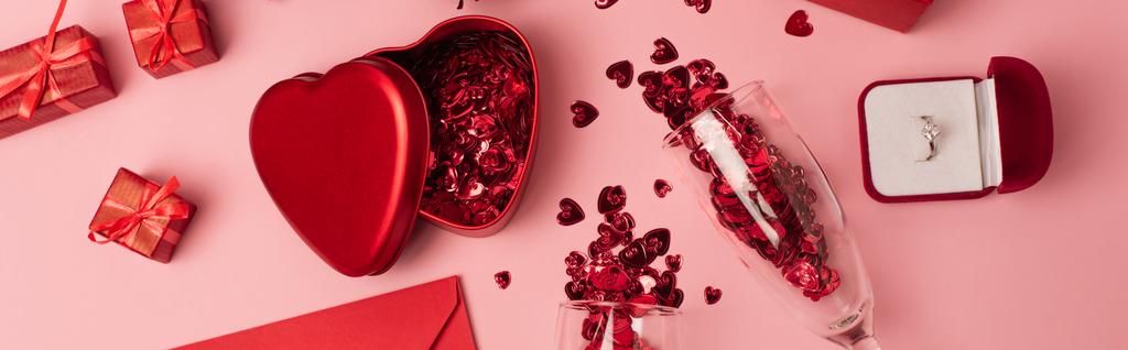 vista superior de copas de champán con confeti brillante cerca de cajas de regalo, anillo de compromiso y sobre en rosa, bandera - Foto, Imagen