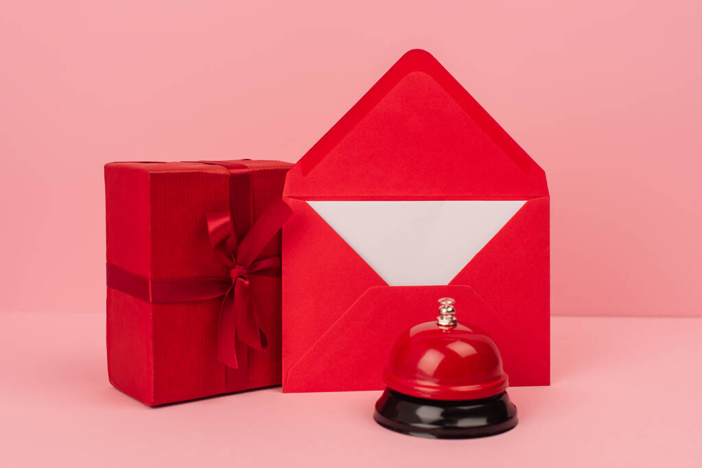 τυλιγμένο κουτί δώρου και κόκκινο φάκελο με γράμμα κοντά σε μεταλλική καμπάνα σε ροζ  - Φωτογραφία, εικόνα