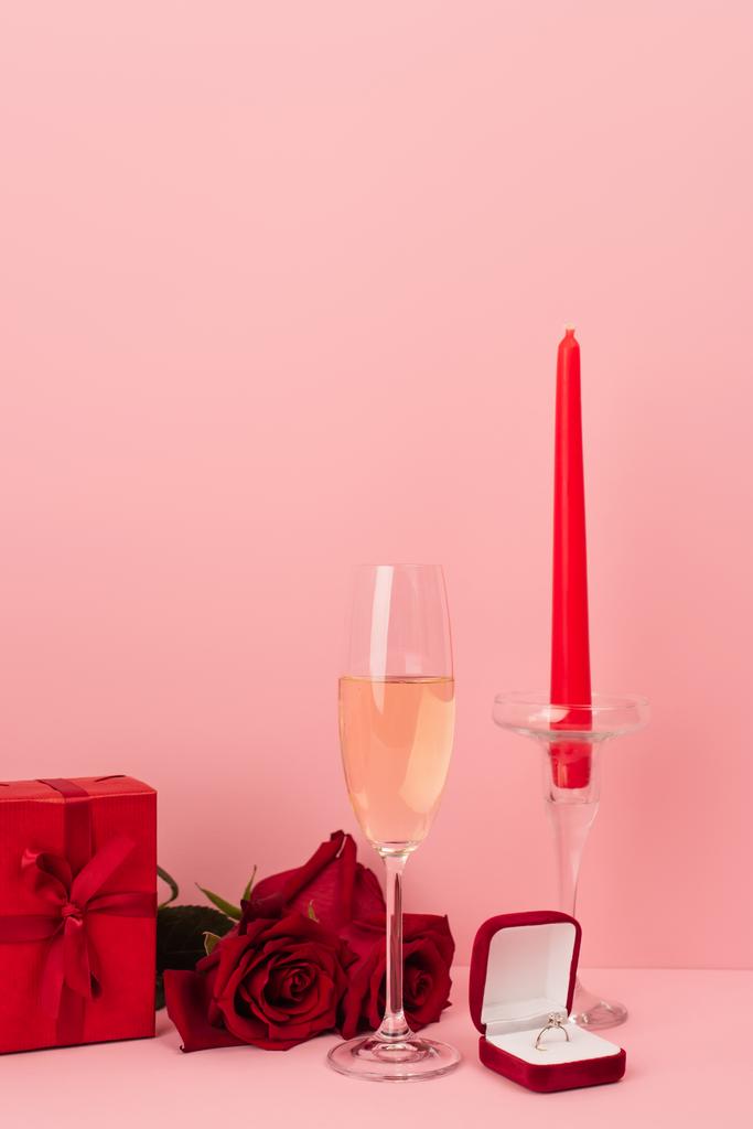 Glas Champagner in der Nähe von Geschenkbox, Rosen, Verlobungsring und Kerze auf rosa  - Foto, Bild