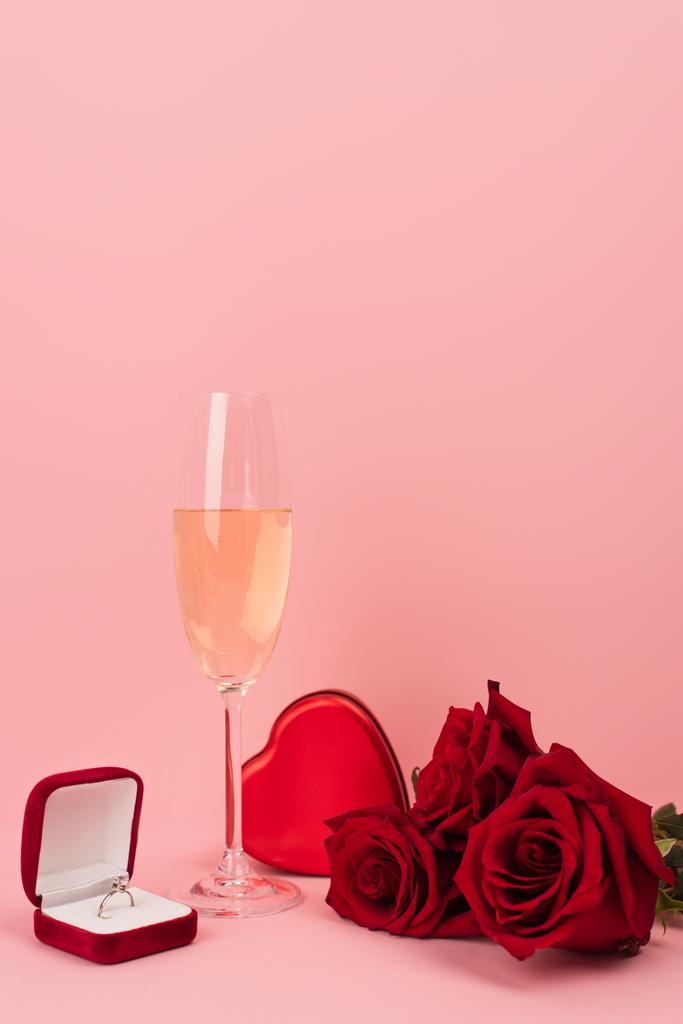 Ένα ποτήρι σαμπάνια κοντά σε κουτί σε σχήμα καρδιάς, τριαντάφυλλα και δαχτυλίδι αρραβώνων σε ροζ  - Φωτογραφία, εικόνα
