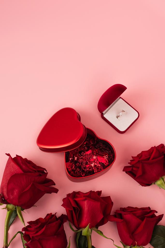 κορυφαία άποψη των κόκκινων τριαντάφυλλων κοντά στο κουτί με κονφετί σε σχήμα καρδιάς και δαχτυλίδι αρραβώνων σε ροζ  - Φωτογραφία, εικόνα