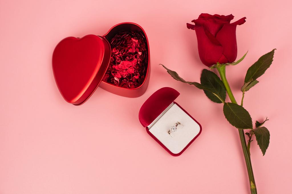 Kutunun yanındaki kırmızı gülün üstünde kalp şeklinde konfeti ve pembe nişan yüzüğü var.  - Fotoğraf, Görsel