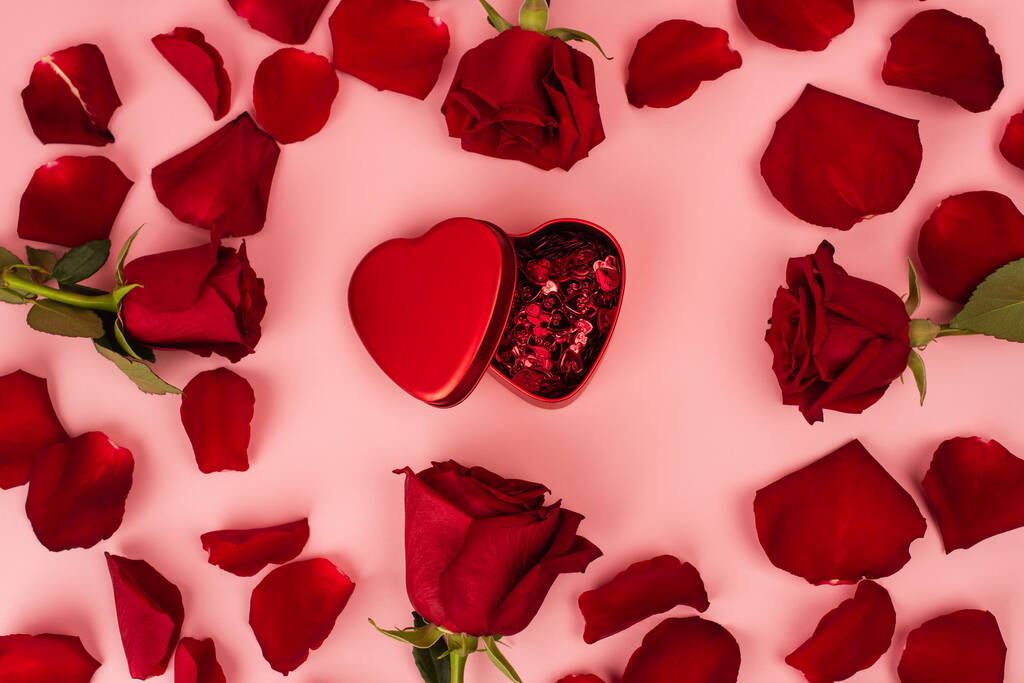 плоские лепестки красной розы вокруг металлической коробки с конфетти на розовый  - Фото, изображение