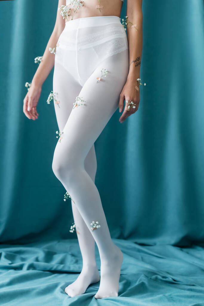 частичный вид татуированной женщины с крошечными цветами на колготках, стоящих на зеленой драпировке - Фото, изображение