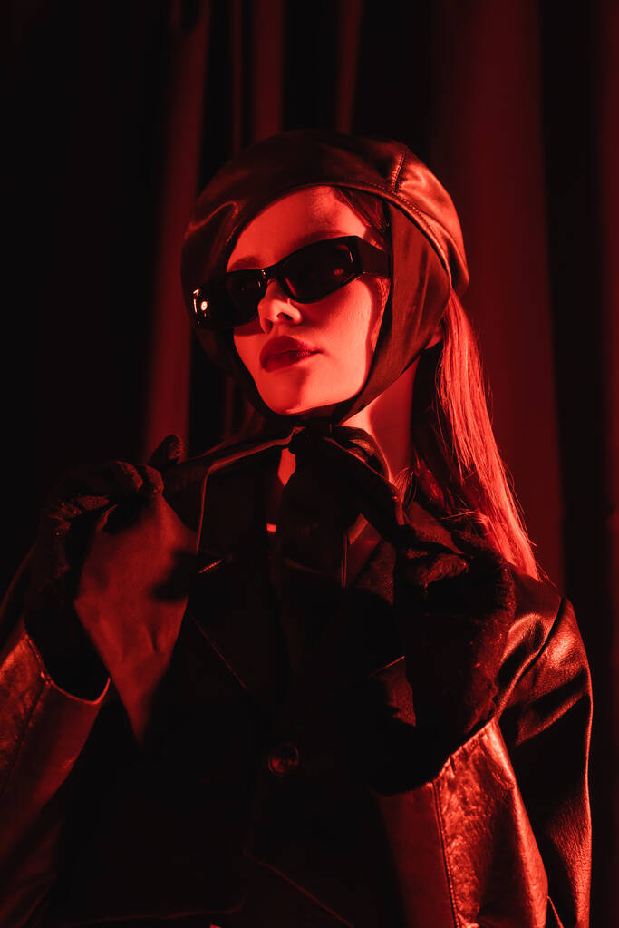 vista de ángulo bajo de la mujer glamour en gafas de sol negras, boina de cuero y guantes de pie sobre fondo oscuro con luz roja  - Foto, Imagen
