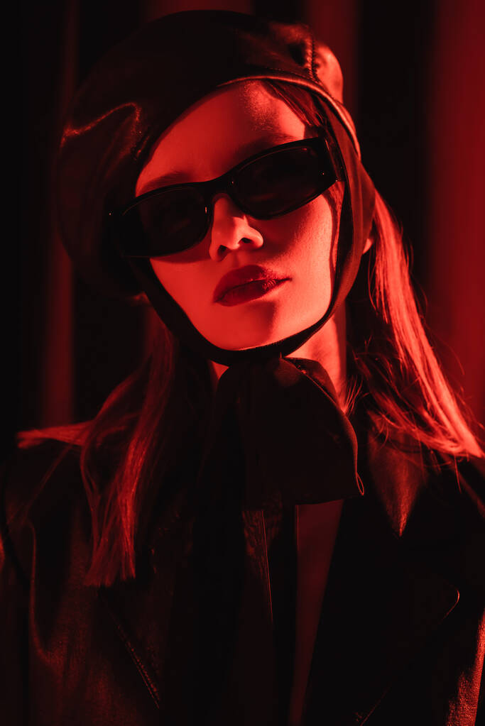Glamour-Frau mit Baskenmütze und schwarzer Sonnenbrille posiert auf dunklem Hintergrund mit rotem Licht - Foto, Bild