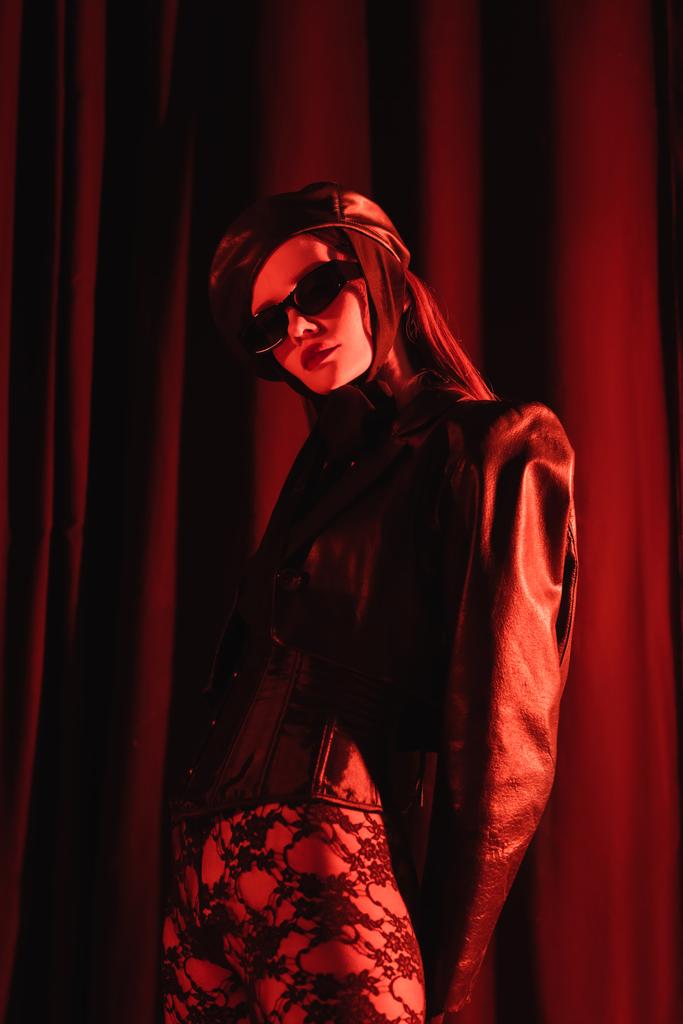 низкий угол зрения сексуальной женщины в кружевных леггинсах, черная кожаная куртка и солнцезащитные очки на темном драпированном фоне  - Фото, изображение