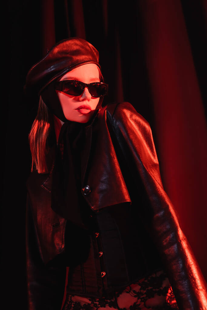 sexy woman in black leather jacket, corset, sunglasses and beret on dark draped background  - Photo, Image