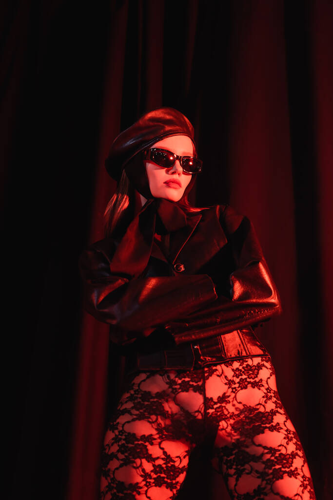 низкий угол зрения на стильную соблазнительную женщину в кружевных леггинсах, кожаной куртке и черных солнцезащитных очках на темном фоне с драпировкой - Фото, изображение