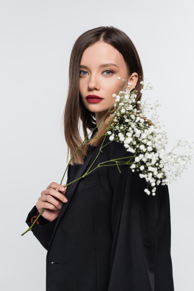 seductive brunette woman in black blazer looking at camera while holding branch of gypsophila isolated on grey - Photo, Image