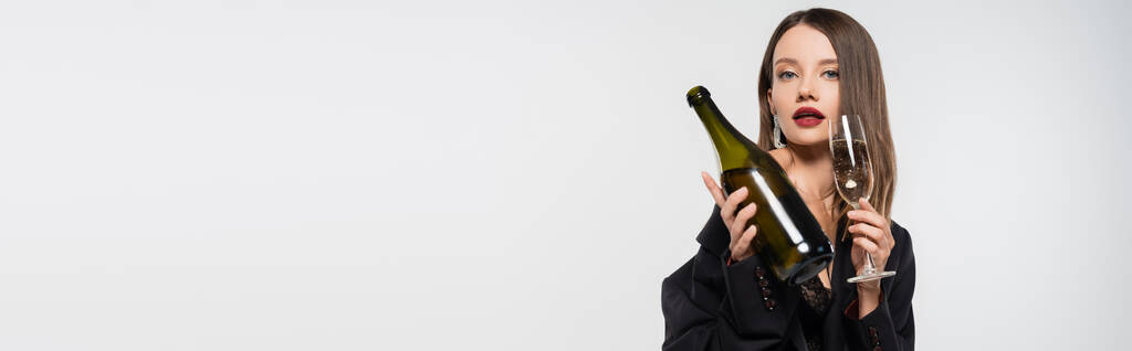 zmysłowa kobieta w czarnej marynarce trzymająca butelkę i kieliszek szampana patrząc na kamerę odizolowaną na szarym, banerze - Zdjęcie, obraz