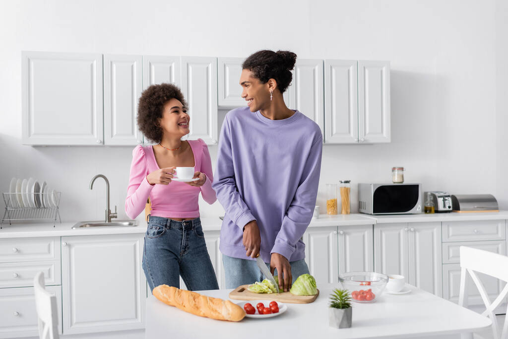 Happy african american woman with cup looking at boyfriend cooking salad in kitchen  - Photo, Image