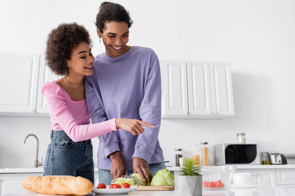Pozitív afro-amerikai nő mutat cseresznye paradicsom, míg a barátja főzés saláta otthon  - Fotó, kép
