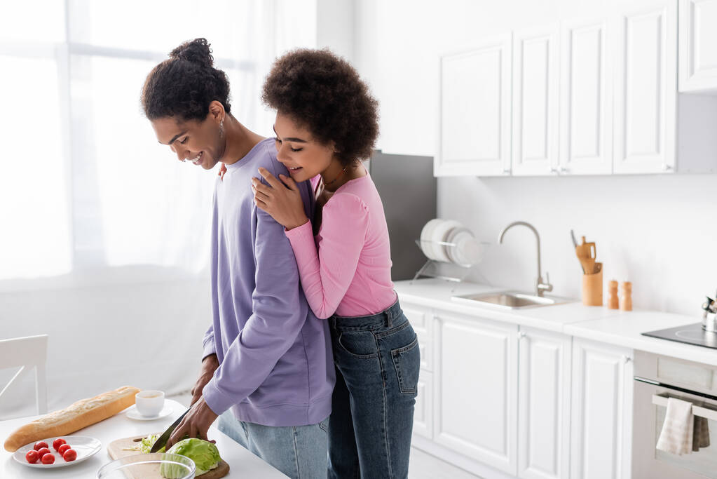 jong afrikaans amerikaans vrouw knuffelen vriend koken salade in de buurt van baguette en koffie in keuken  - Foto, afbeelding