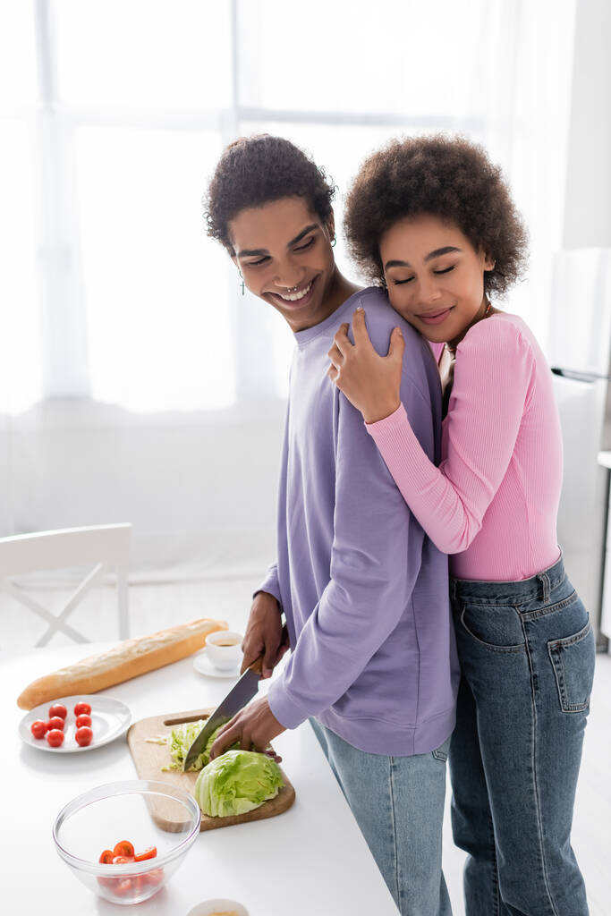 Young african american woman hugging boyfriend cooking salad at home  - Photo, Image