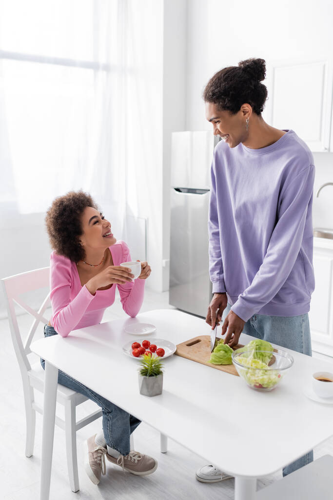Glückliche Afroamerikanerin hält Tasse in der Hand und schaut Freund beim Salatkochen in der Küche an  - Foto, Bild
