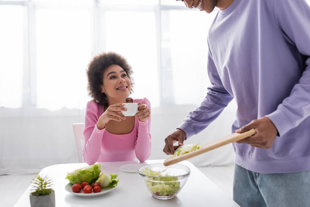 Jeune femme afro-américaine tenant une tasse près de petit ami cuisine salade dans la cuisine  - Photo, image