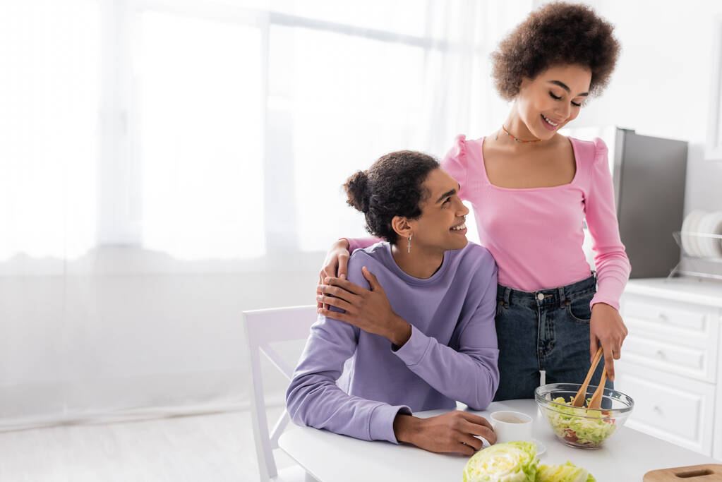 アフリカ系アメリカ人女性抱擁ボーイフレンドと調理サラダでキッチン  - 写真・画像