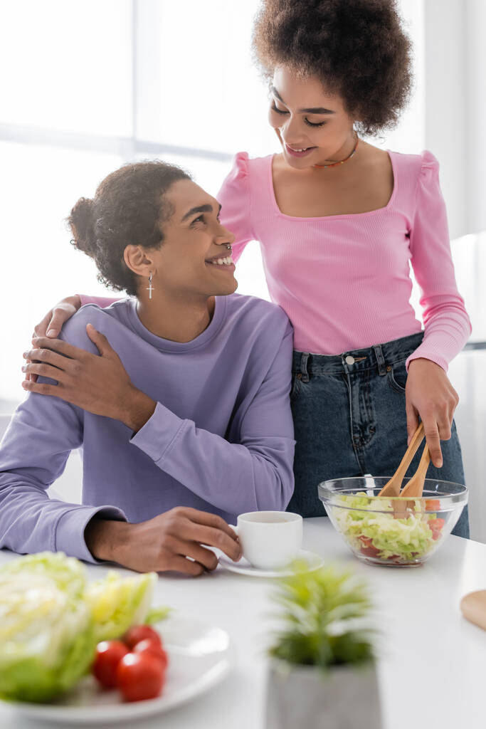 Χαμογελώντας αφροαμερικανή γυναίκα αγκαλιάζει τον φίλο της με το κύπελλο, ενώ η μαγειρική σαλάτα στην κουζίνα  - Φωτογραφία, εικόνα