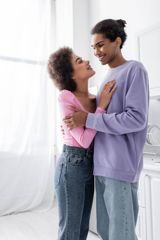 Positiv afrikanisch-amerikanisches Paar schaut einander an, während es sich zu Hause umarmt  - Foto, Bild