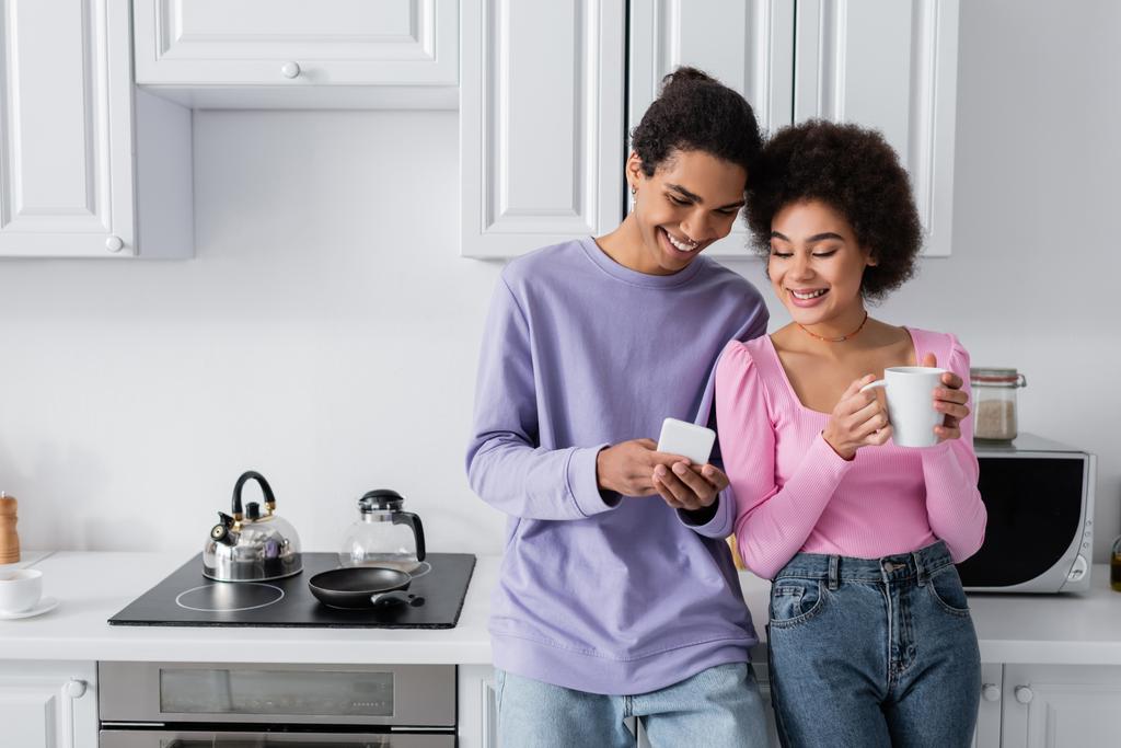 正アフリカ系アメリカ人男性保持スマートフォン近くガールフレンドとともにカップでキッチン  - 写真・画像