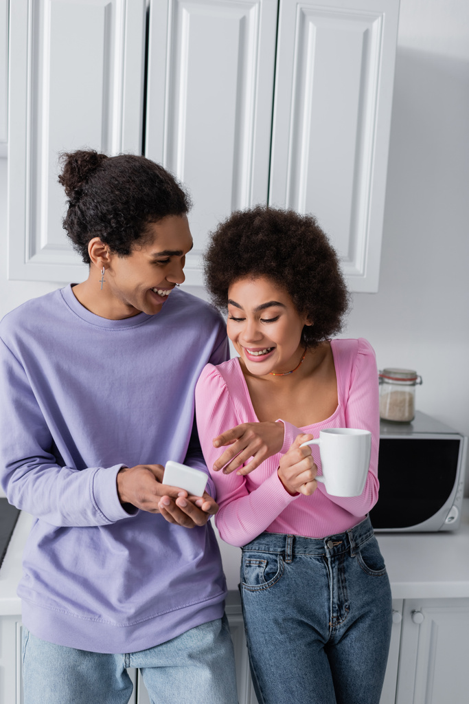 African american woman with cup pointing at smartphone near boyfriend at home  - Photo, Image