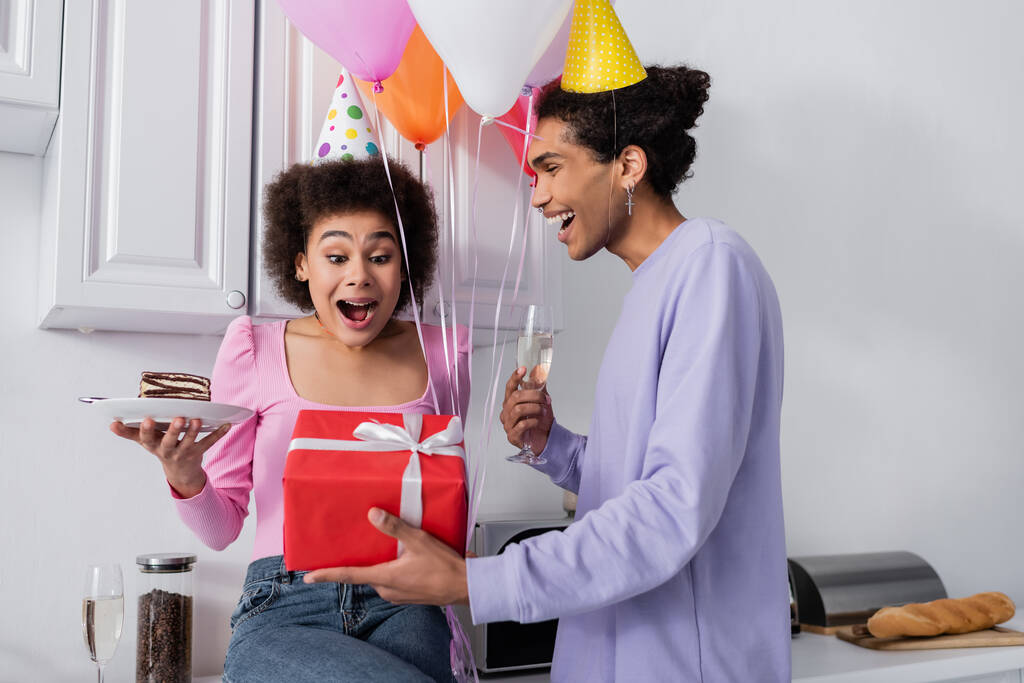 Захоплена афро-американська жінка тримає торт на день народження поруч з хлопцем з шампанським і подарунок на кухні.  - Фото, зображення