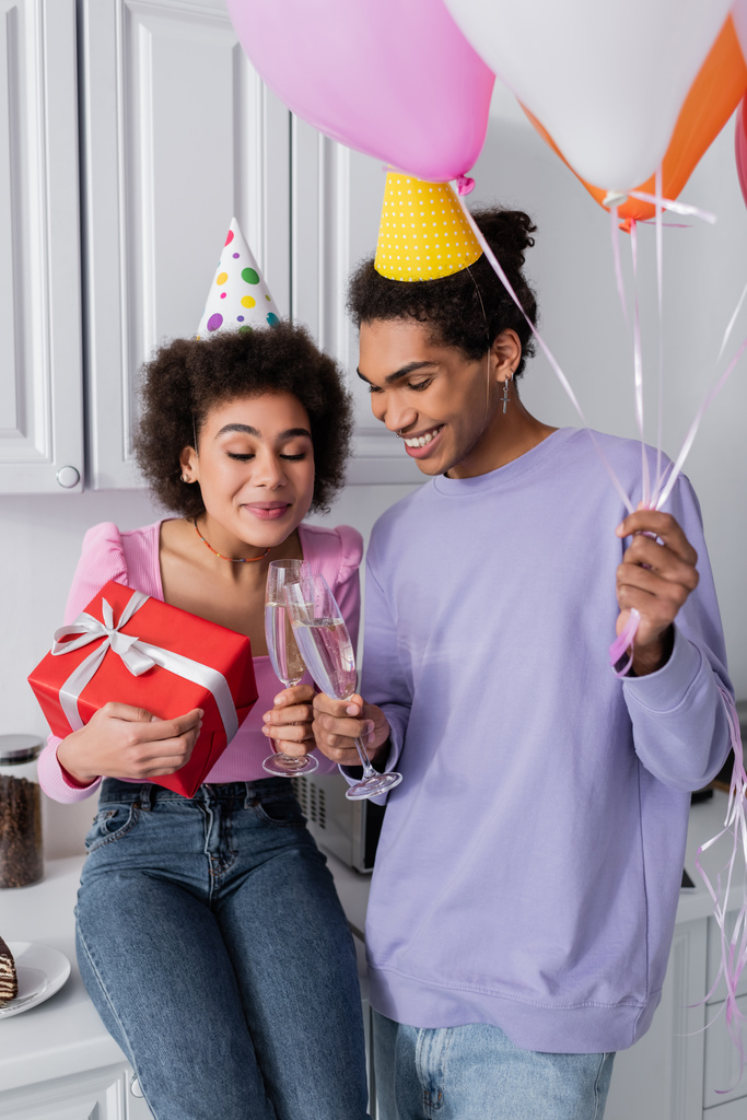 Усміхнений афроамериканець тримає повітряні кулі і чіпляється за шампанське з подругою, яка тримає подарунок на кухні.   - Фото, зображення