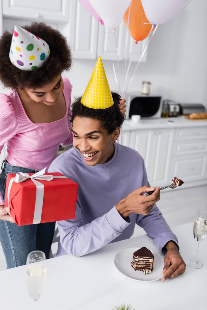 Positif homme afro-américain tenant gâteau d'anniversaire près de petite amie avec cadeau et ballons dans la cuisine  - Photo, image