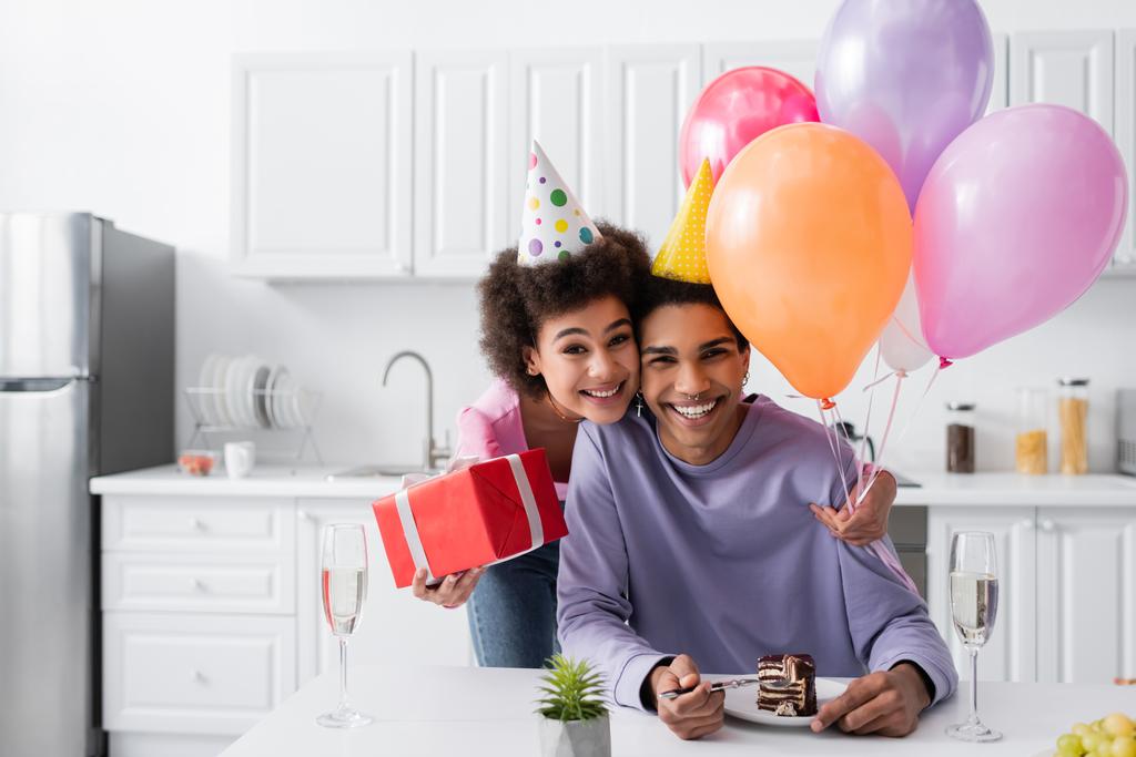 Feliz mujer afroamericana en gorra de fiesta sosteniendo regalo y globos cerca de novio, pastel de cumpleaños y champán en casa  - Foto, Imagen