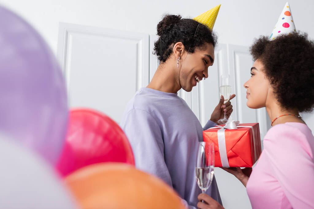 Vue latérale de la femme afro-américaine tenant champagne et cadeau près du petit ami dans le chapeau de fête et des ballons flous à la maison  - Photo, image