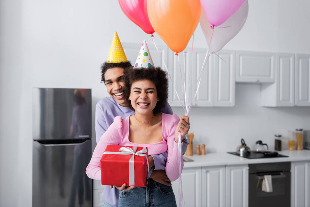 Jeune homme afro-américain en bonnet de fête étreignant petite amie avec cadeau et ballons à la maison  - Photo, image