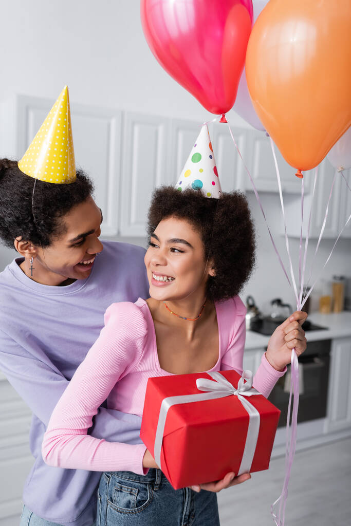 Smiling african american man in party cap embracing girlfriend with festive balloons and gift at home  - Photo, Image