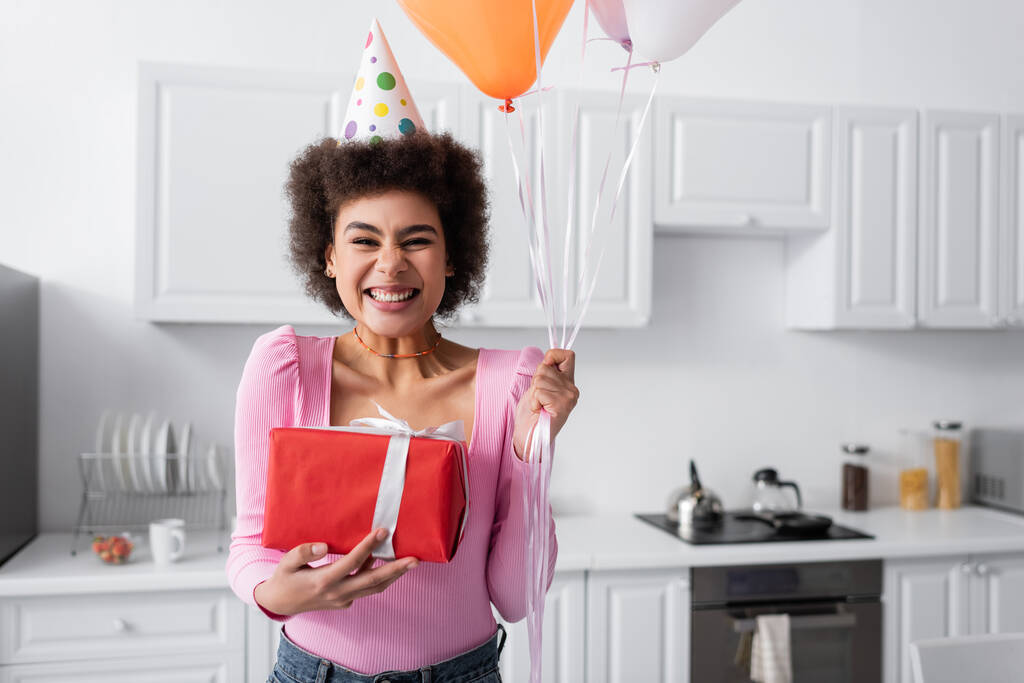 Vrolijk Afrikaans amerikaanse vrouw in party cap houden geschenk doos en ballonnen thuis  - Foto, afbeelding