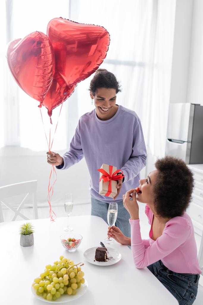 Sonriente hombre afroamericano sosteniendo globos en forma de corazón y regalo cerca de la novia con fresa y champán en casa  - Foto, imagen