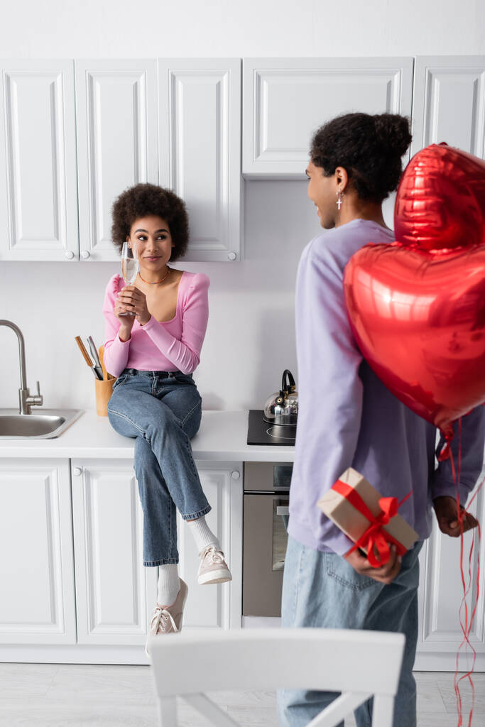 African american man hiding heart-shaped balloons and present near girlfriend with champagne at home  - Foto, Imagem