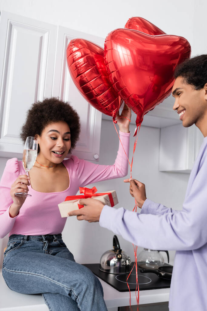 Smiling african american woman holding champagne near boyfriend with heart-shaped balloons and gift box in kitchen  - Foto, Bild