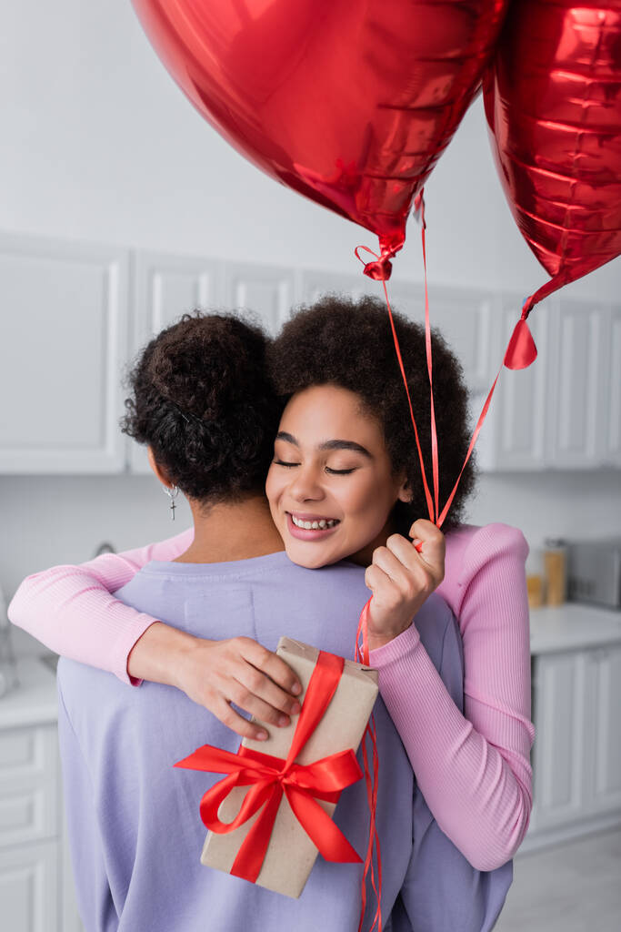 Νεαρή Αφροαμερικανή γυναίκα που κρατάει μπαλόνια σε σχήμα καρδιάς και δώρο ενώ αγκαλιάζει το αγόρι της στο σπίτι  - Φωτογραφία, εικόνα