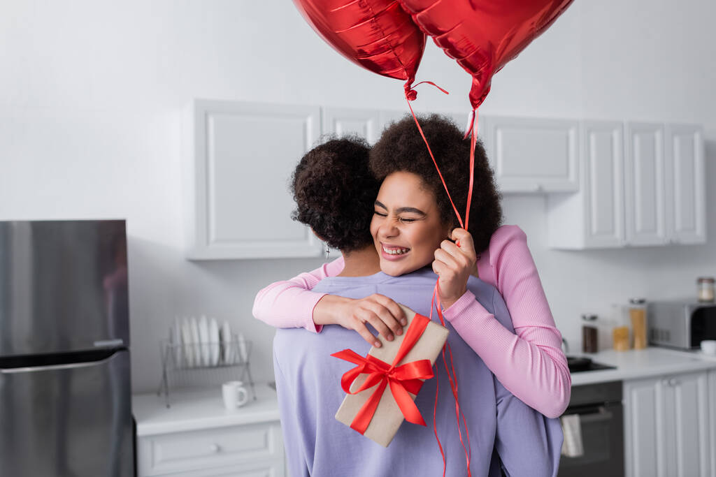 Femme afro-américaine joyeuse tenant des ballons et un cadeau tout en embrassant petit ami à la maison  - Photo, image