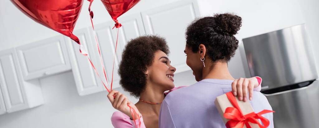 Smiling african american woman holding balloons and gift box while embracing boyfriend in kitchen, banner  - Foto, Imagem