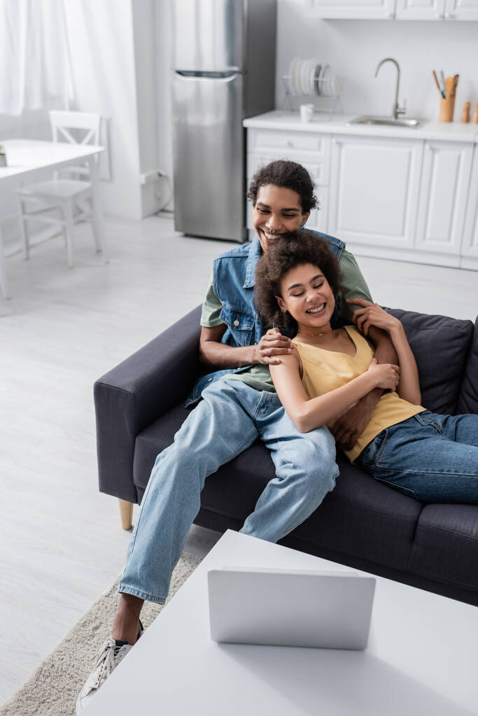 African american man hugging smiling girlfriend and looking at laptop at home  - Foto, Bild