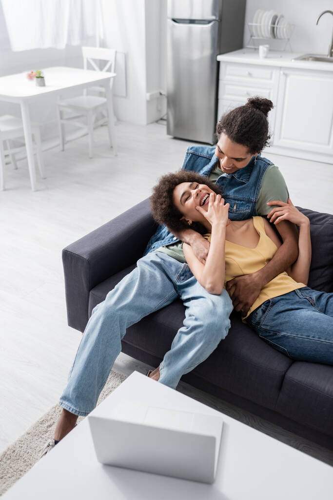 homme afro-américain positif étreignant petite amie joyeuse sur le canapé près d'un ordinateur portable à la maison  - Photo, image
