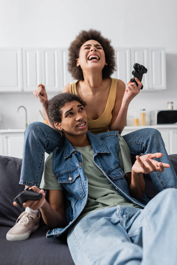 KYIV, UCRANIA - 9 de noviembre de 2021: Emocionada mujer afroamericana sosteniendo un joystick cerca de su novio disgustado en el sofá  - Foto, imagen
