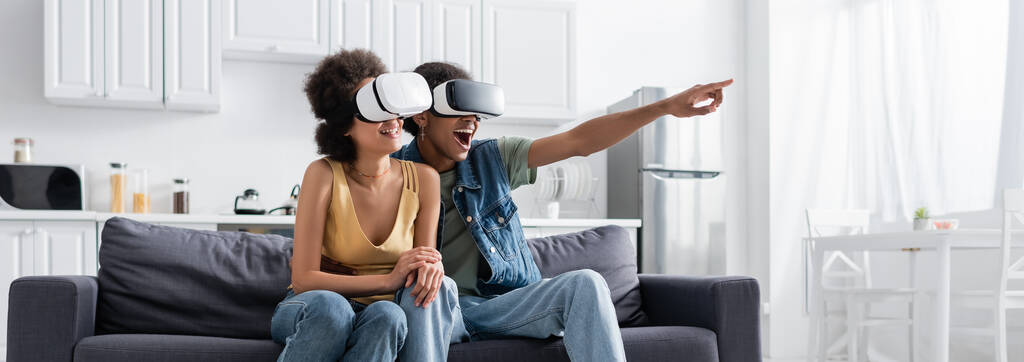 Heyecanlı Afro-Amerikan adam, VR kulaklıklı, parmaklarıyla evde gülümseyen kız arkadaşını işaret ediyor.  - Fotoğraf, Görsel