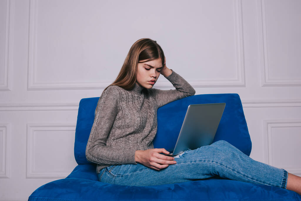 Kazaklı, kot pantolonlu, uzaktan kumandalı bir kadın netbook 'u karıştırıyor ve beyaz duvara karşı kanepede otururken projeyi düşünüyor. - Fotoğraf, Görsel