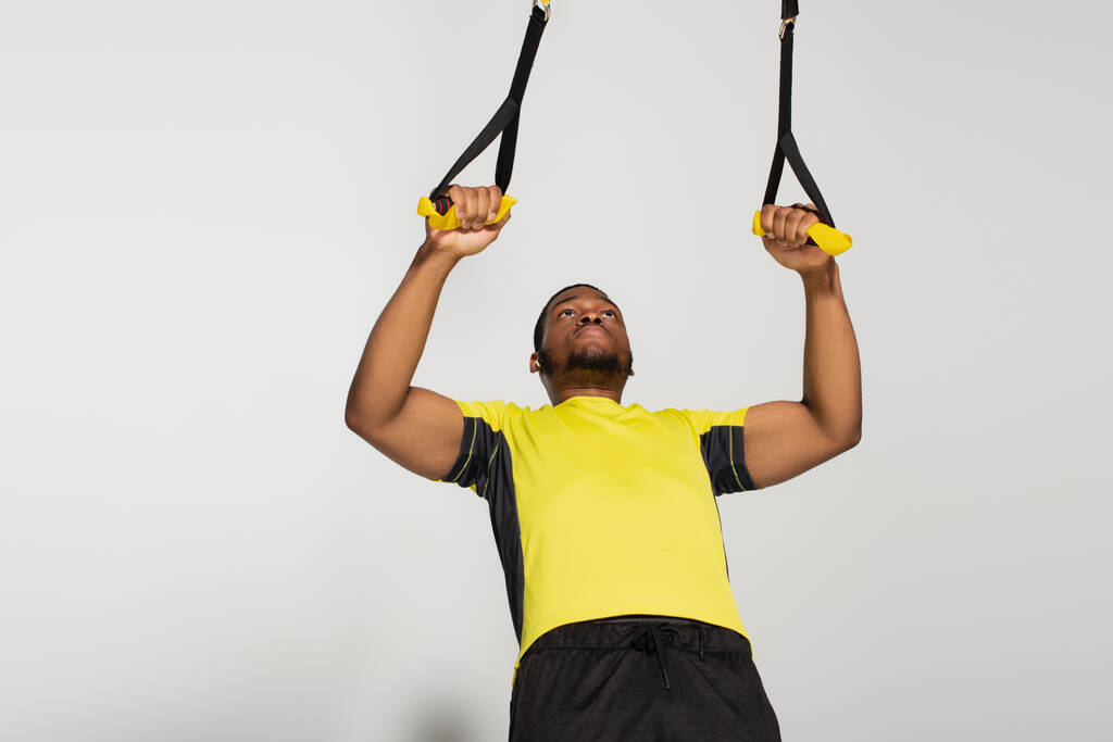 μυώδης Αφροαμερικάνος αθλητής που γυμνάζεται με ζώνες αντίστασης στο γκρι - Φωτογραφία, εικόνα