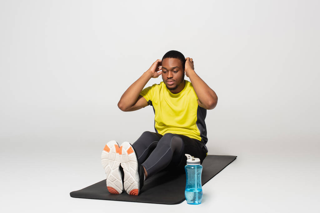 longueur totale de sportif afro-américain faisant de l'exercice abs sur tapis de fitness près de bouteille de sport sur gris - Photo, image