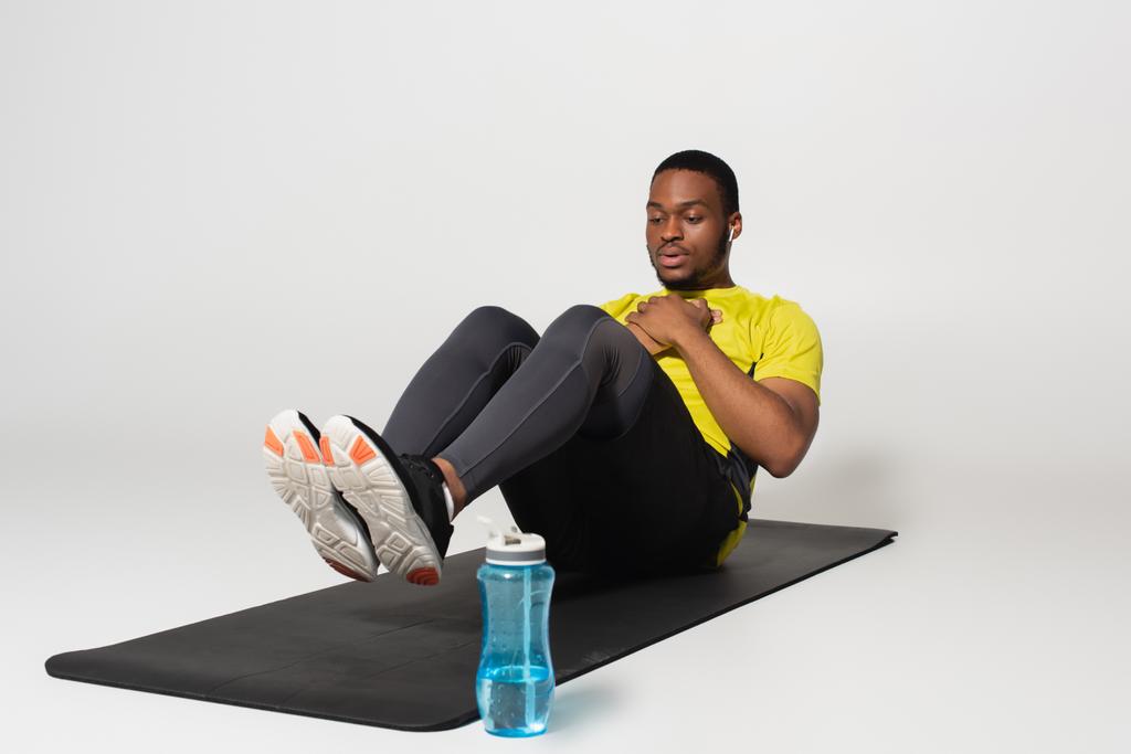 volledige lengte van sportieve Afro-Amerikaanse man in oortelefoons doet buikspieroefeningen op fitness mat in de buurt van sportfles op grijs - Foto, afbeelding
