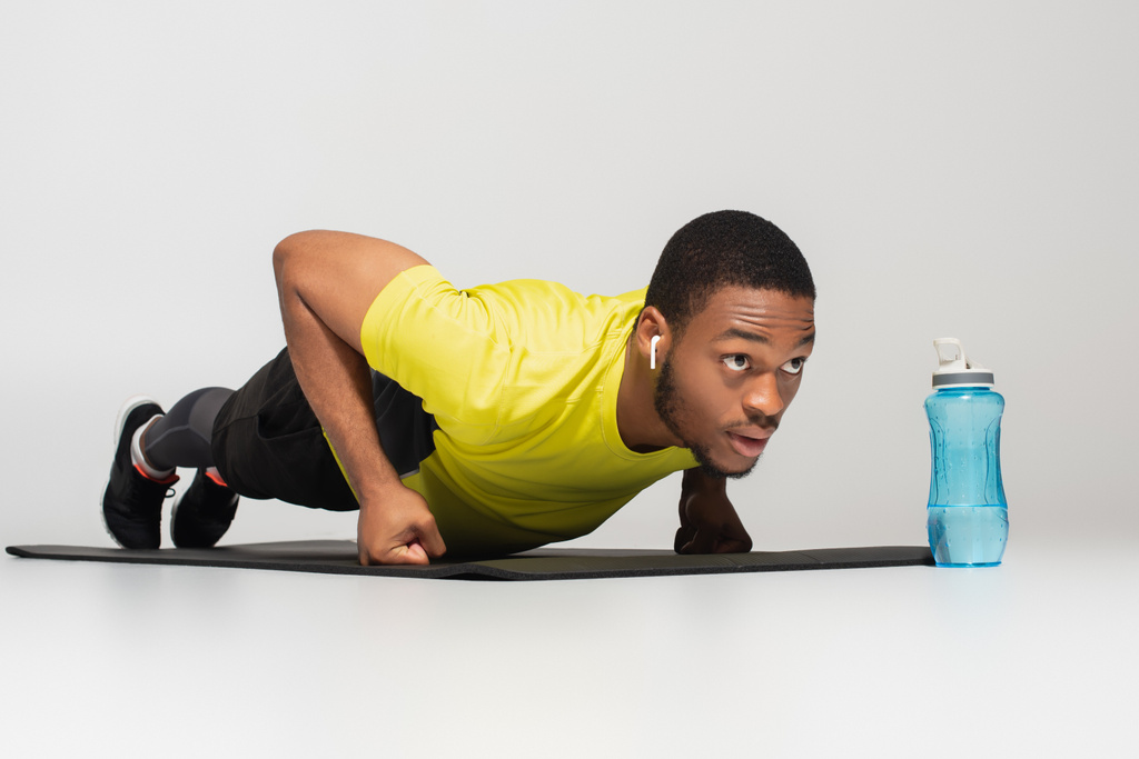 спортивный африканский американец в наушниках делает отжимания на фитнес-коврик возле спортивной бутылки, изолированной на сером - Фото, изображение